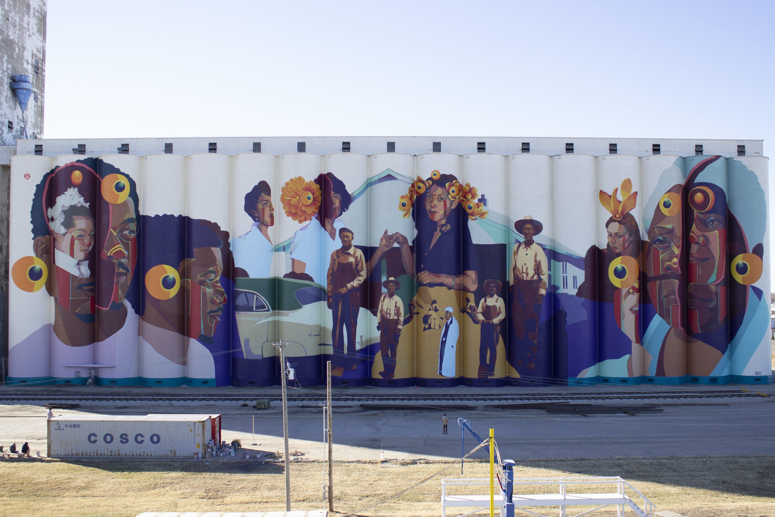 A huge mural is painted on the side of a grain elevator, featuring a variety of men and women of many backgrounds.